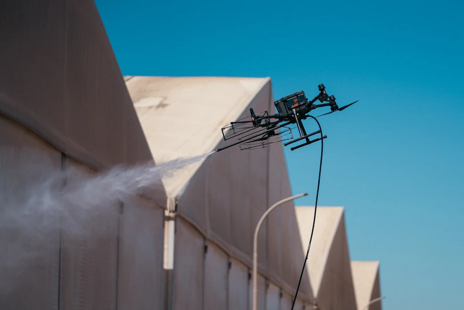 Dron de AGS lavando con agua a presión la fachada de una nave industrial, con la manguera de alimentación colgando bajo el aparato