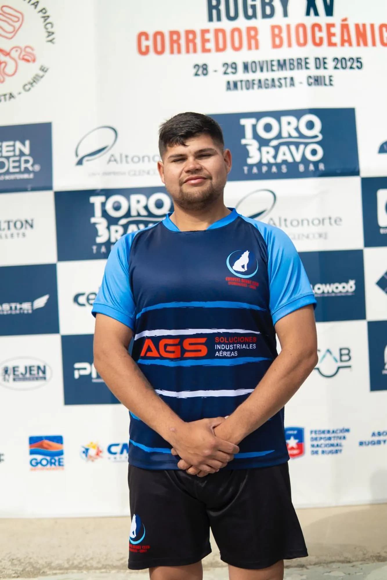 Un jugador de Coyotes Antofagasta con la camiseta auspiciada por AGS, ante el panel de auspiciadores del torneo Rugby XV Corredor Bioceánico de Antofagasta